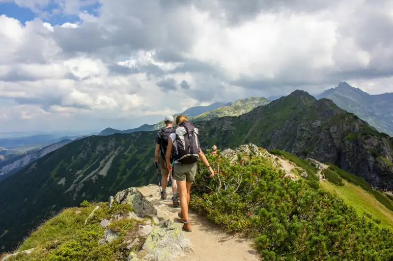 A man and a woman hiking a trail at the top of a mountain, with more mountains and a cloudy sky in the background.