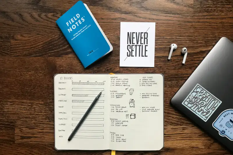A laptop, ear buds, notebook, and a small sign that reads 'Never settle' all sitting on a desk.